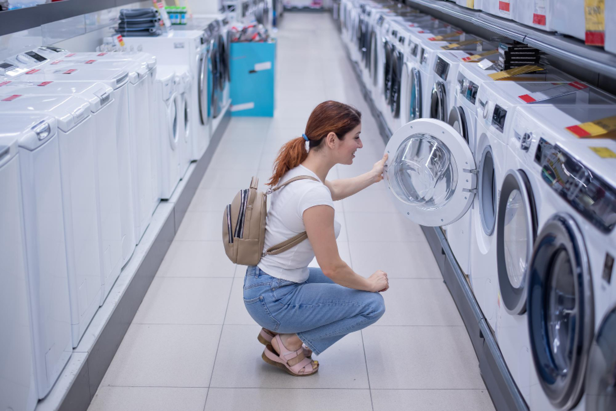 Mulher agachada inspecionando o tambor de uma máquina de lavar de abertura frontal (Front Load) em uma loja, representando a escolha da melhor máquina de lavar sem agitador.